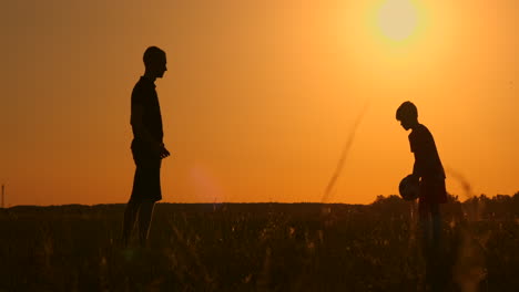 Vater-Und-Sohn-Spielen-Fußball-Im-Park-Bei-Sonnenuntergang,-Silhouetten-Vor-Dem-Hintergrund-Einer-Zeitlupenaufnahme-Einer-Hellen-Sonne.